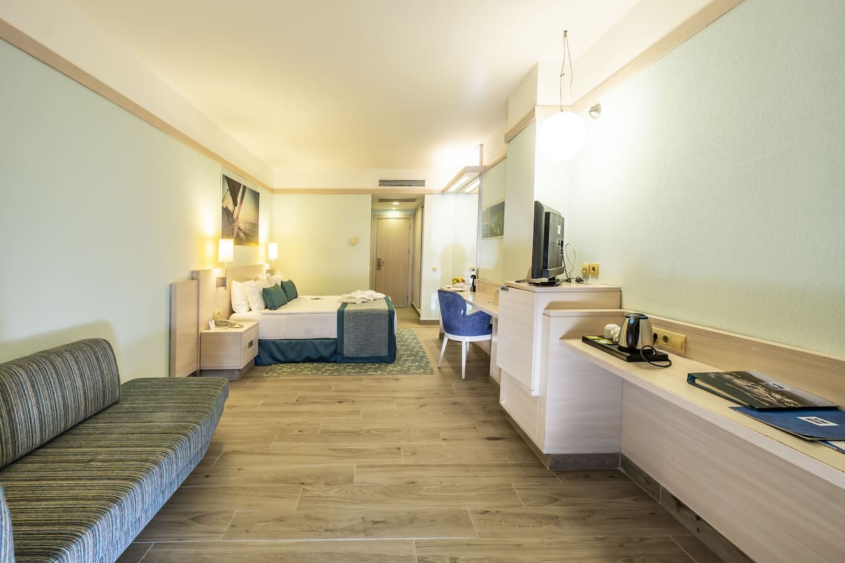 Quarto de hotel com cama, escrivaninha, poltrona, sofá e televisão.