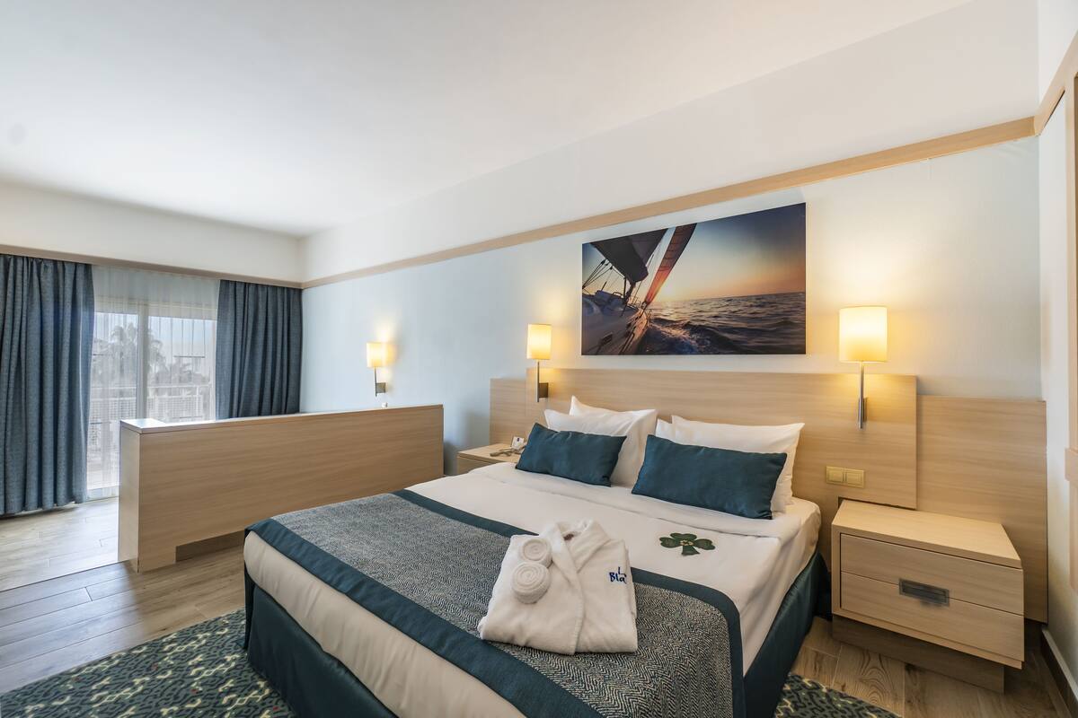 Quarto de hotel com uma cama grande, uma escrivaninha, duas mesas de cabeceira, um quadro na parede e vista para a cidade pela janela.