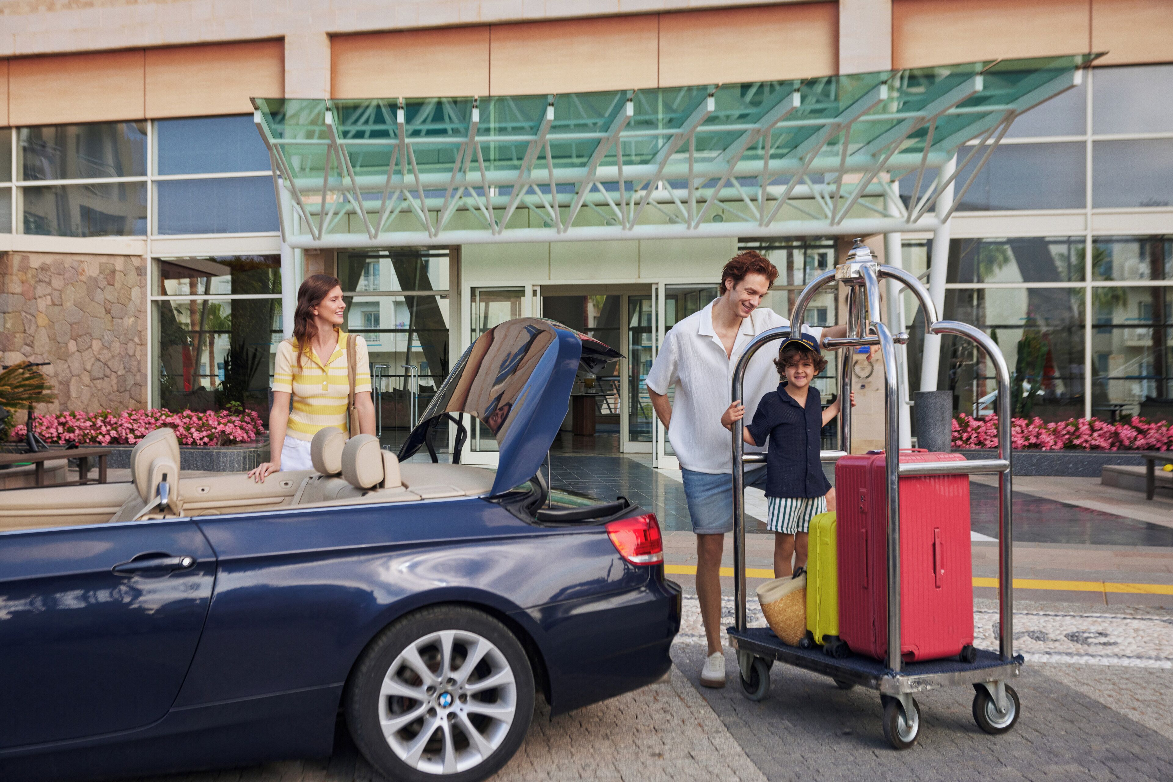 A hotel entrance with a glass canopy, a blue convertible car, and a luggage cart with suitcases.