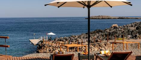 Eine Holzterrasse mit Liegestühlen und einem großen Sonnenschirm mit Blick auf eine felsige Küstenlinie und das Meer.