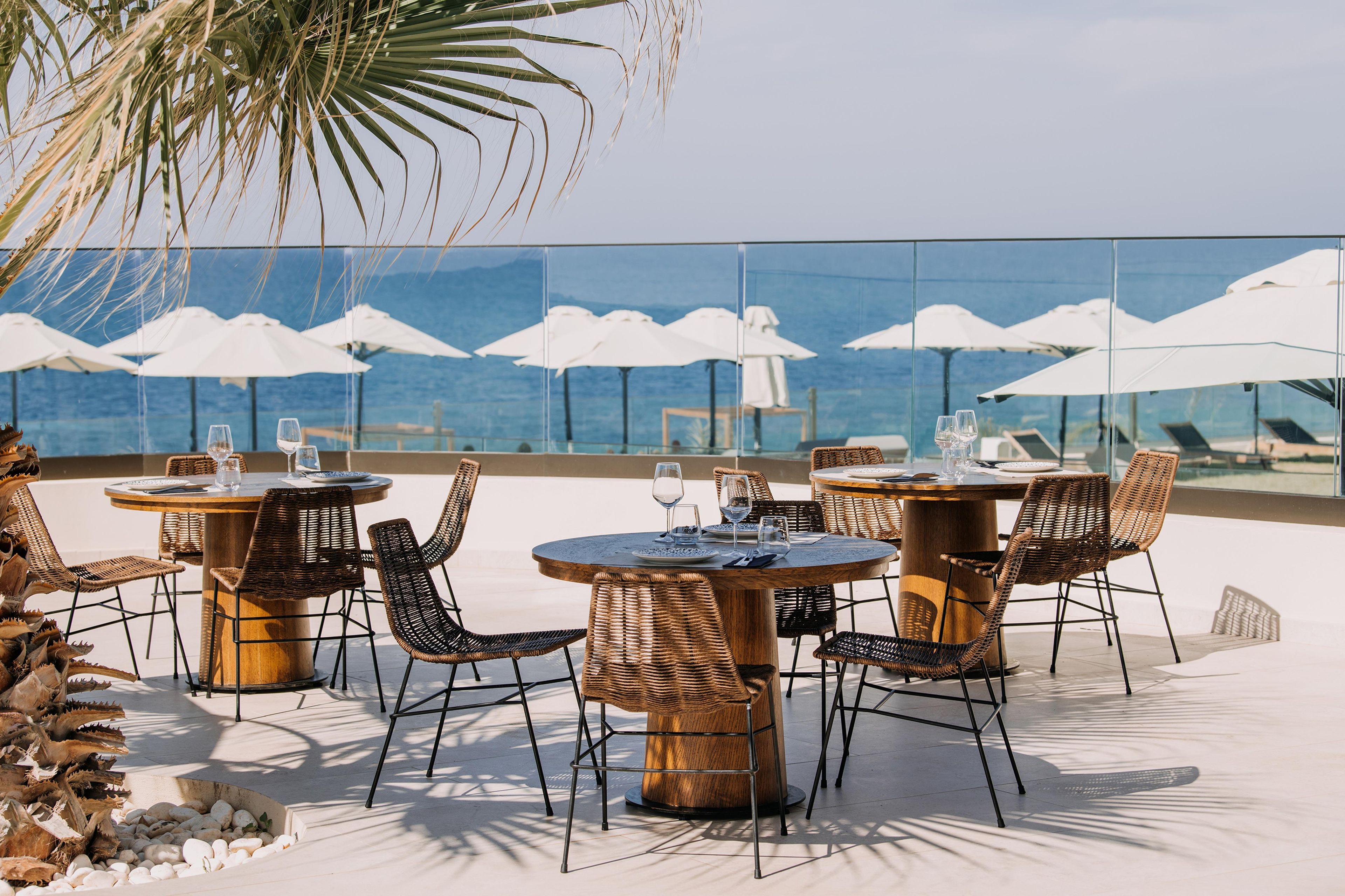 Mesas de comedor al aire libre con sillas de mimbre y sombrillas blancas, con vista a un cuerpo de agua.