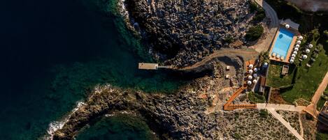 An aerial view of a coastal property with a swimming pool, a rocky shoreline, and a paved pathway.