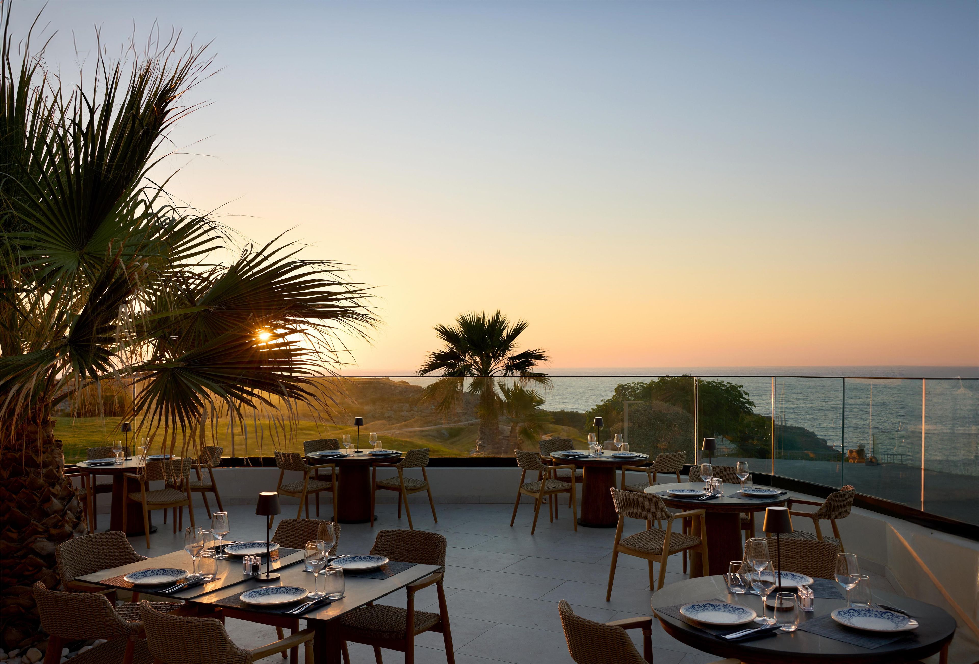 A dining area with tables set for two, overlooking a coastal landscape during sunset.