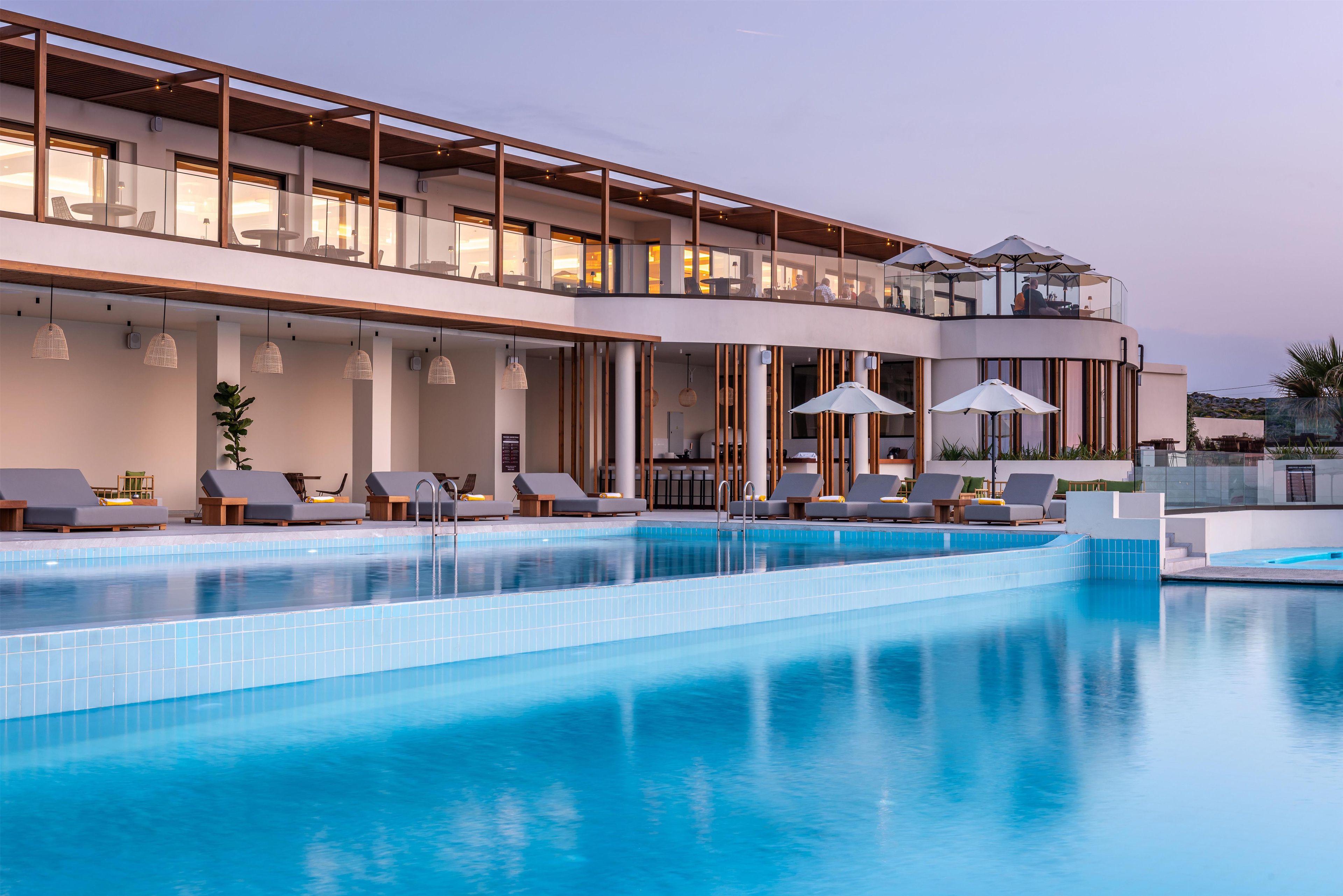Un hotel moderno con una amplia piscina exterior, sillas de descanso y un balcón en el segundo piso con asientos.