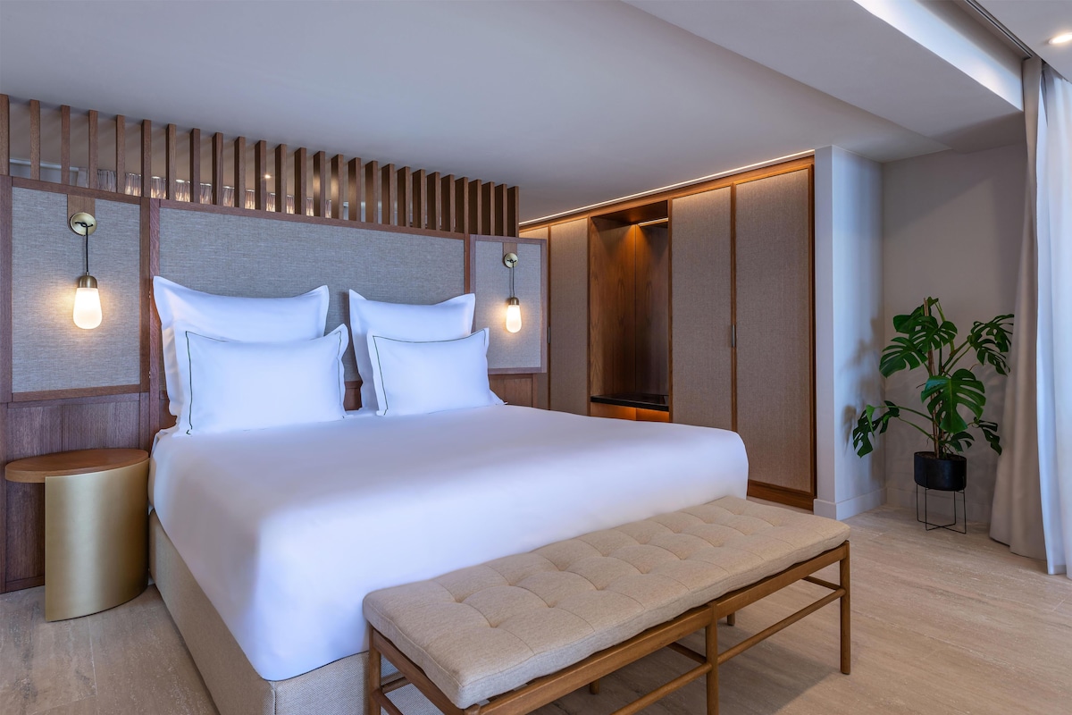 A modern hotel room with a large bed, bedside tables, a bench, and a potted plant.