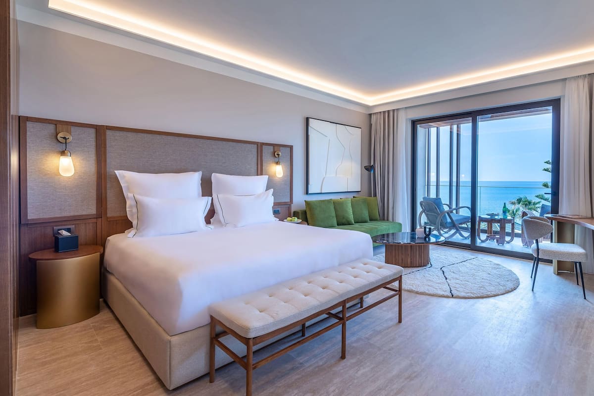 A modern hotel room with a large bed, a green sofa, a dining area with a table and chairs, and a balcony with a view of the sea.