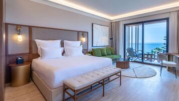 Une chambre d’hôtel moderne dotée d’un grand lit, d’un canapé vert, d’un coin repas avec une table et des chaises, et d’un balcon offrant une vue sur la mer.