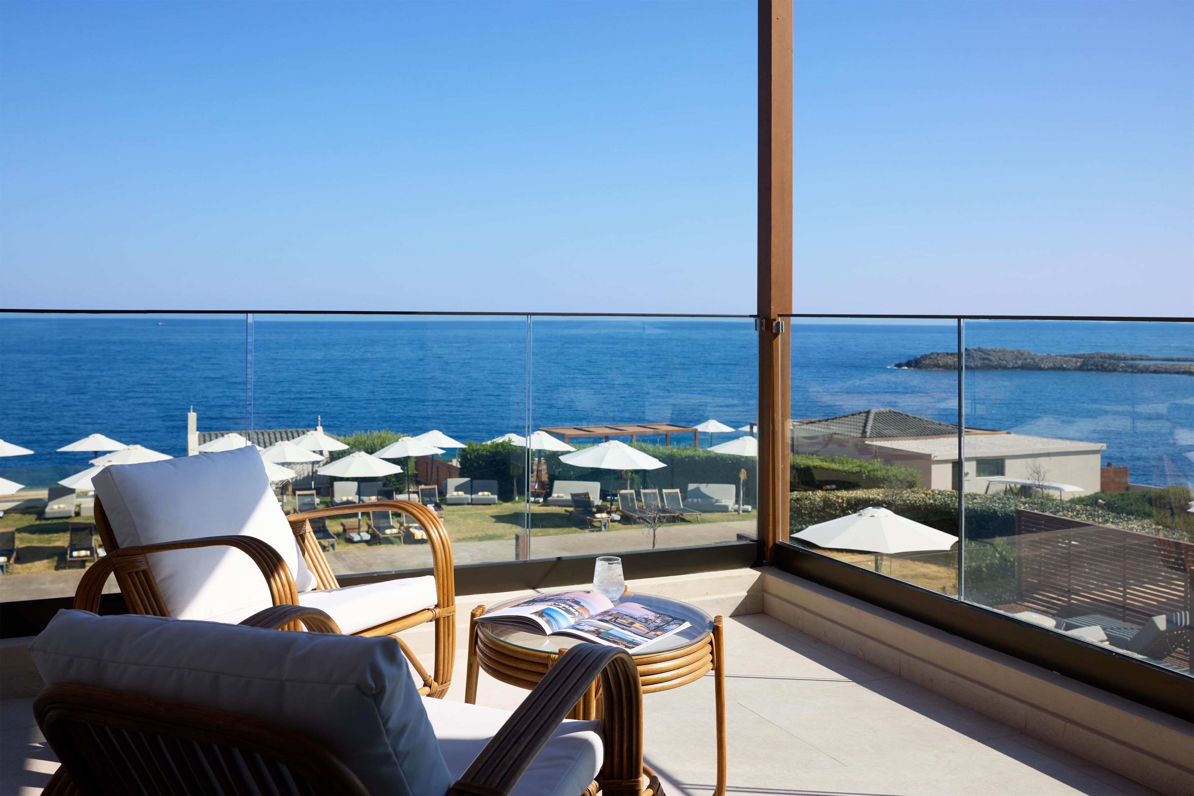 A balcony with wicker chairs and a glass table, overlooking a coastal area with umbrellas and lounge chairs.
