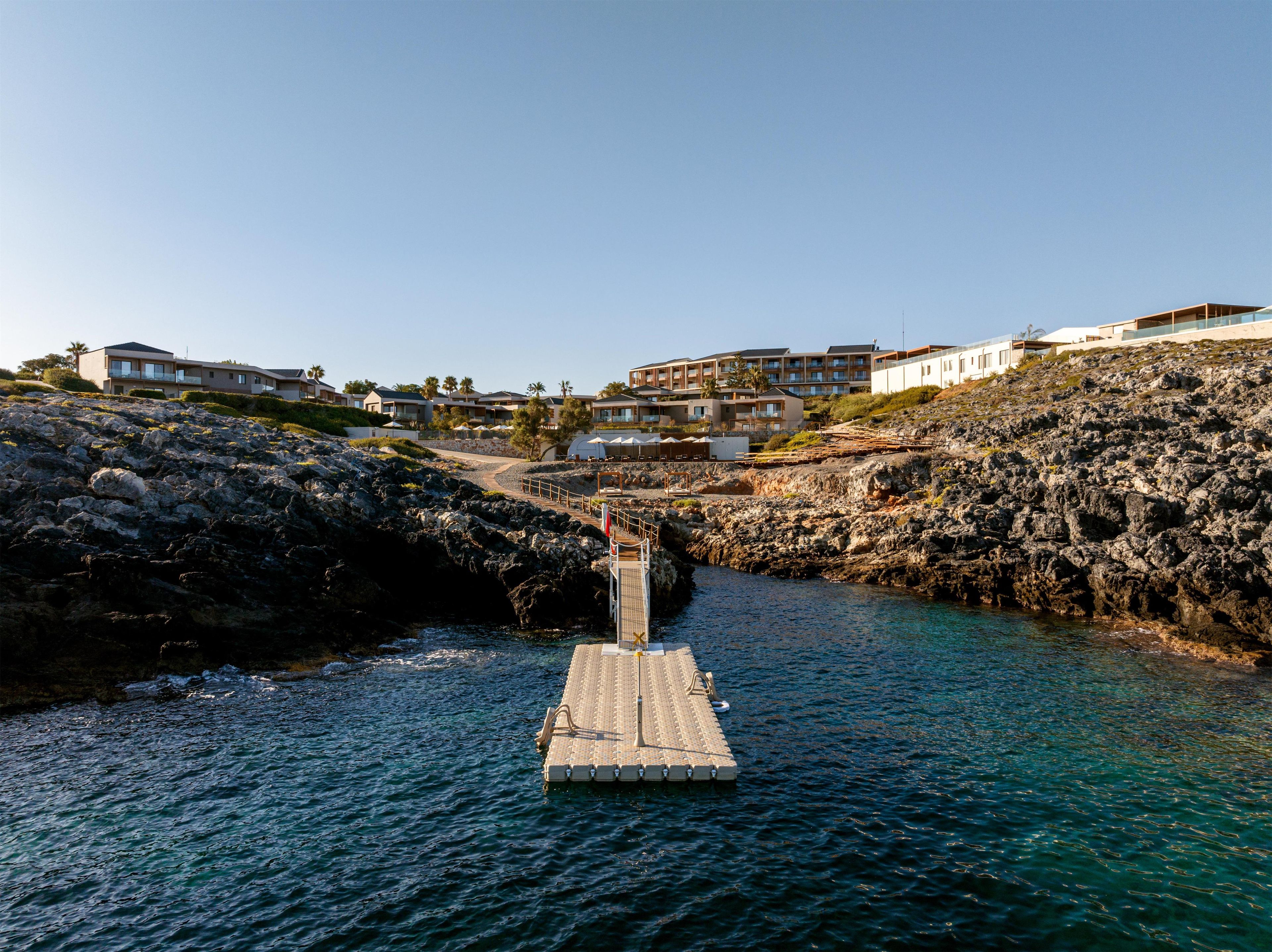 Propiedad costera con muelle que desemboca en una costa rocosa, rodeada de edificios modernos con varios balcones.