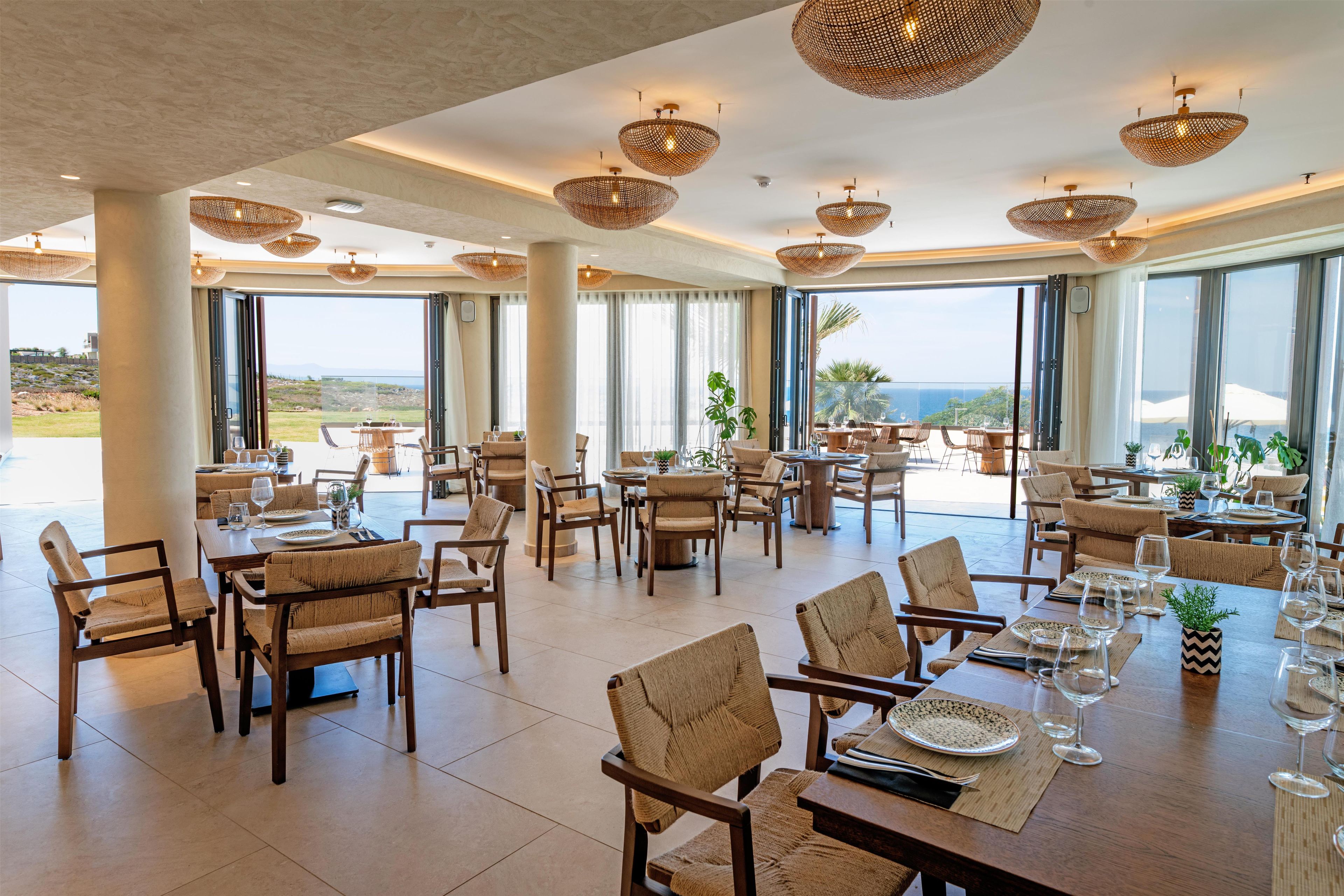 A spacious dining area with high ceilings, large windows, and wicker ceiling lamps. Tables are set with chairs and dining ware.