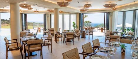 A spacious dining area with high ceilings, large windows, and wicker ceiling lamps. Tables are set with chairs and dining ware.