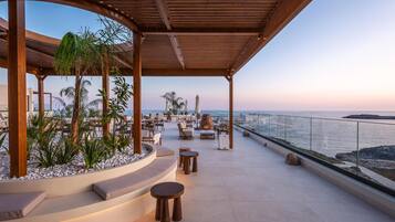 A covered outdoor seating area with a view of the sea, featuring wooden furniture, plants, and a curved seating arrangement.