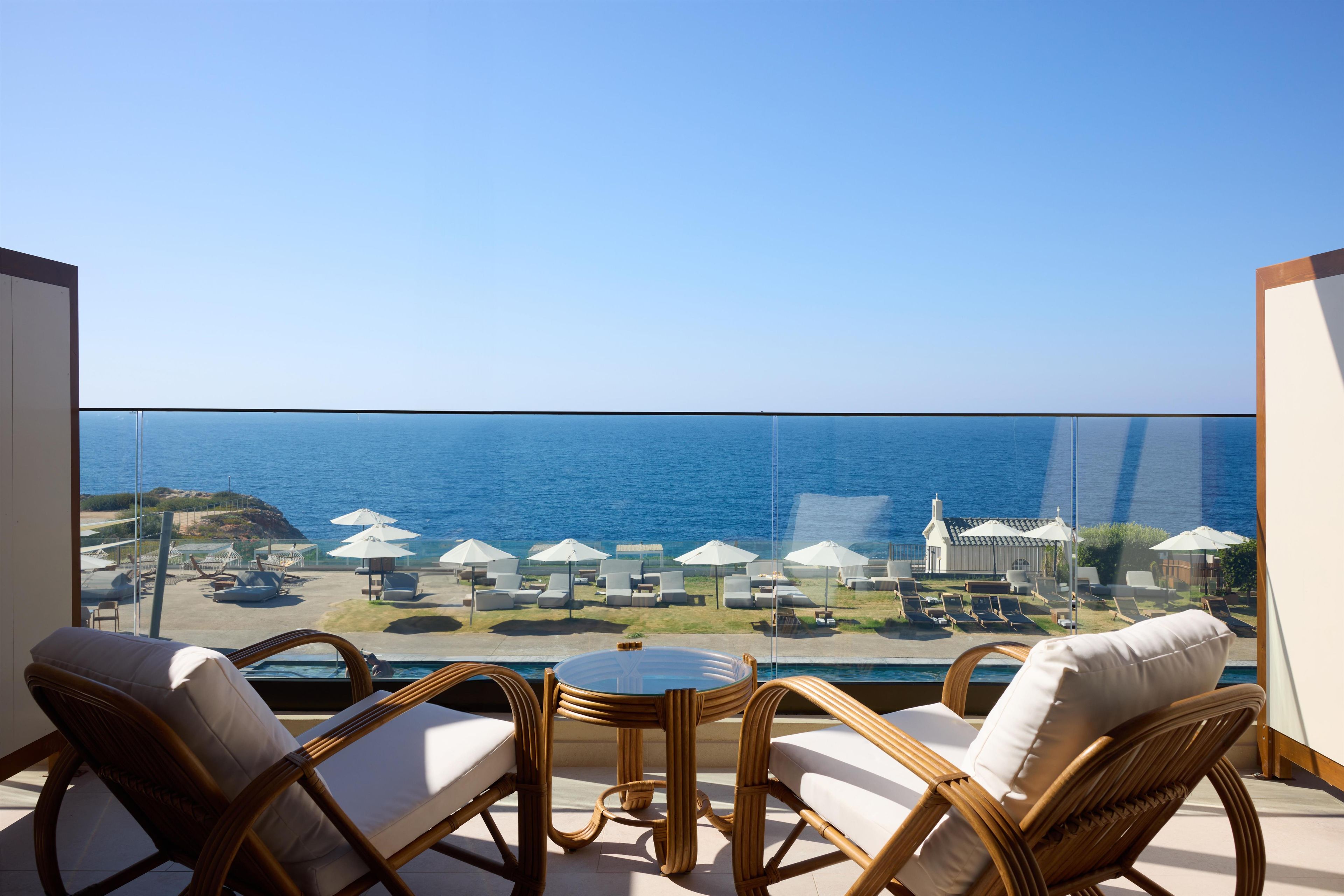 Un balcón con mobiliario de mimbre, una mesa de vidrio y vistas al mar y sombrillas de playa.