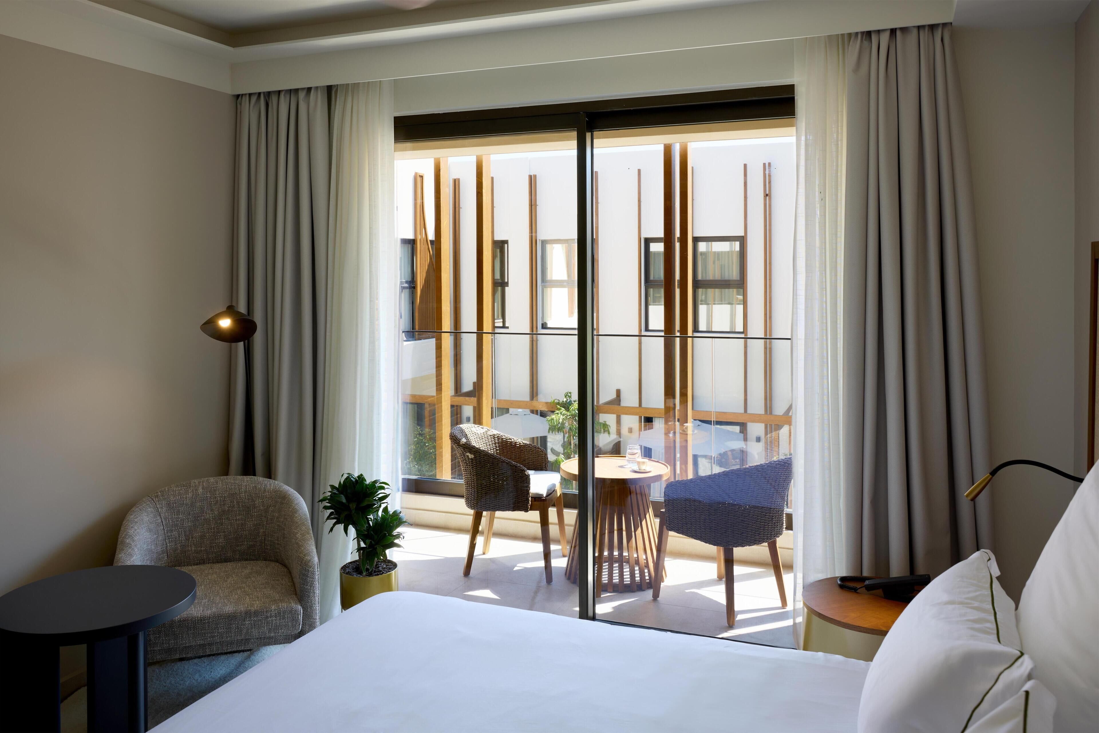 A hotel room with a bed, a small table, a chair, and a balcony with a view of buildings.