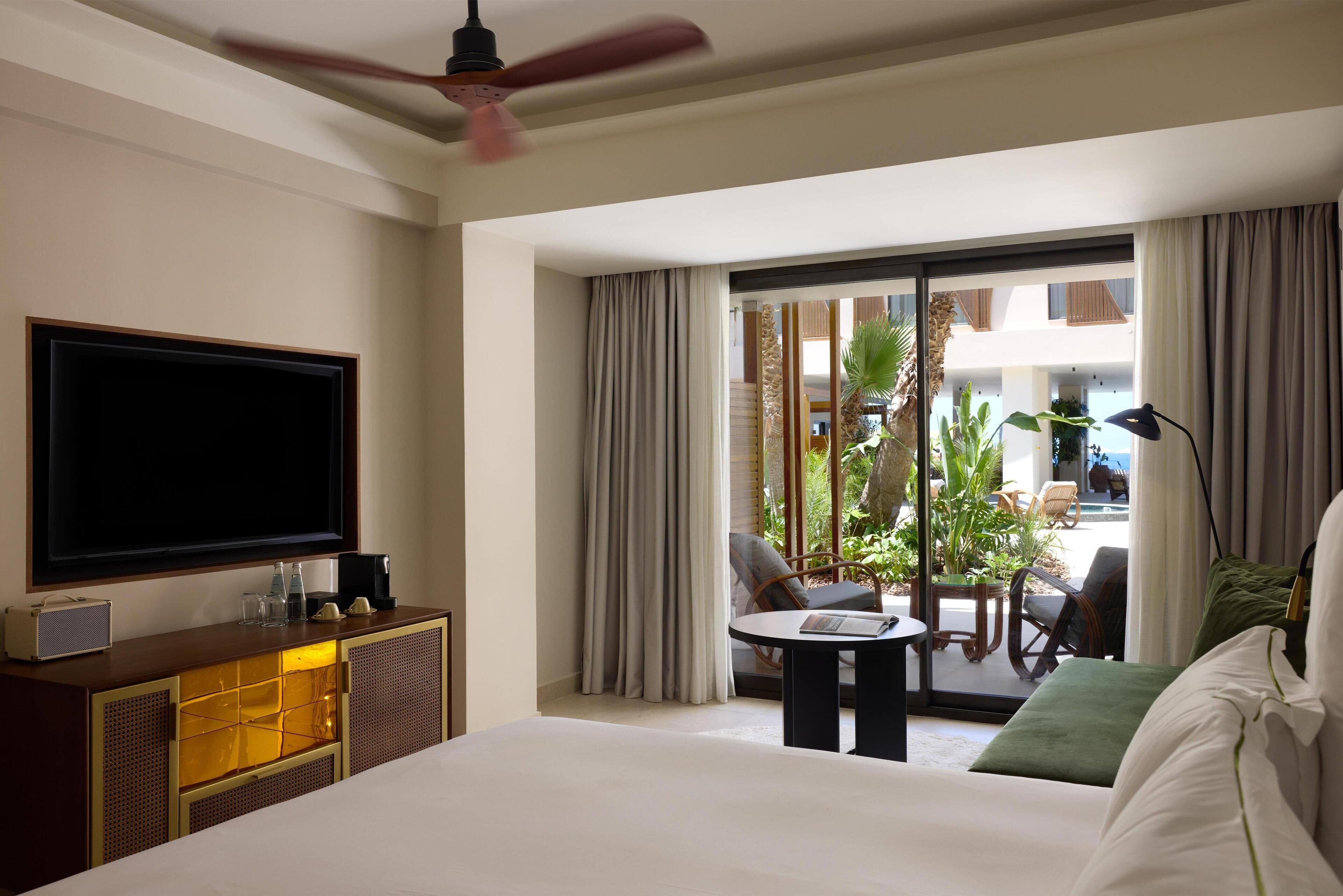 A hotel room with a bed, a television, a desk with a lamp, a balcony with plants and seating, and a ceiling fan.