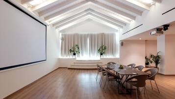 A conference room with a large screen, wooden flooring, a high ceiling with exposed beams, a dining table with chairs, and potted plants.
