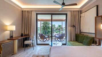 Habitación de hotel con cama, escritorio, silla, una pequeña mesa con bebidas, ventilador de techo y balcón con sillas y vistas a árboles.