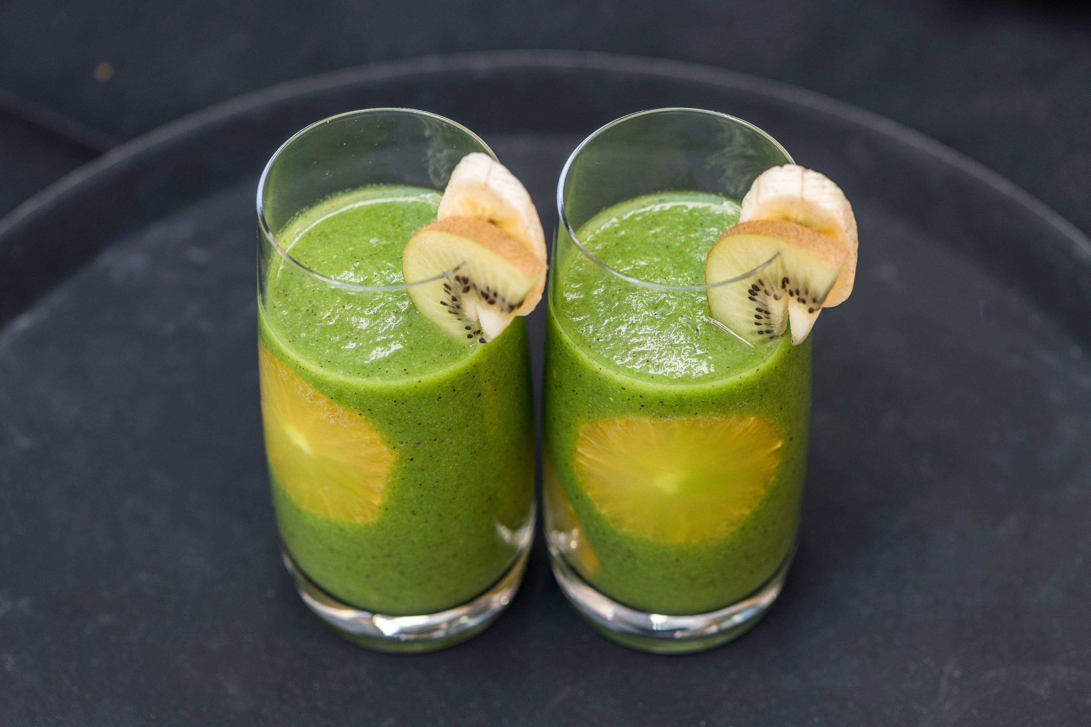 Two glasses of green smoothie with banana slices on a dark surface.