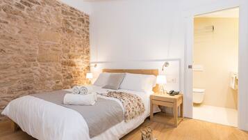 Un dormitorio con pared de piedra, cama de madera, mesita de noche y baño visible a través de una puerta abierta.