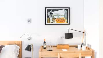 Una habitación de hotel con un escritorio de madera, una cama con ropa blanca, una obra de arte enmarcada en la pared y una mesita de noche con una lámpara.