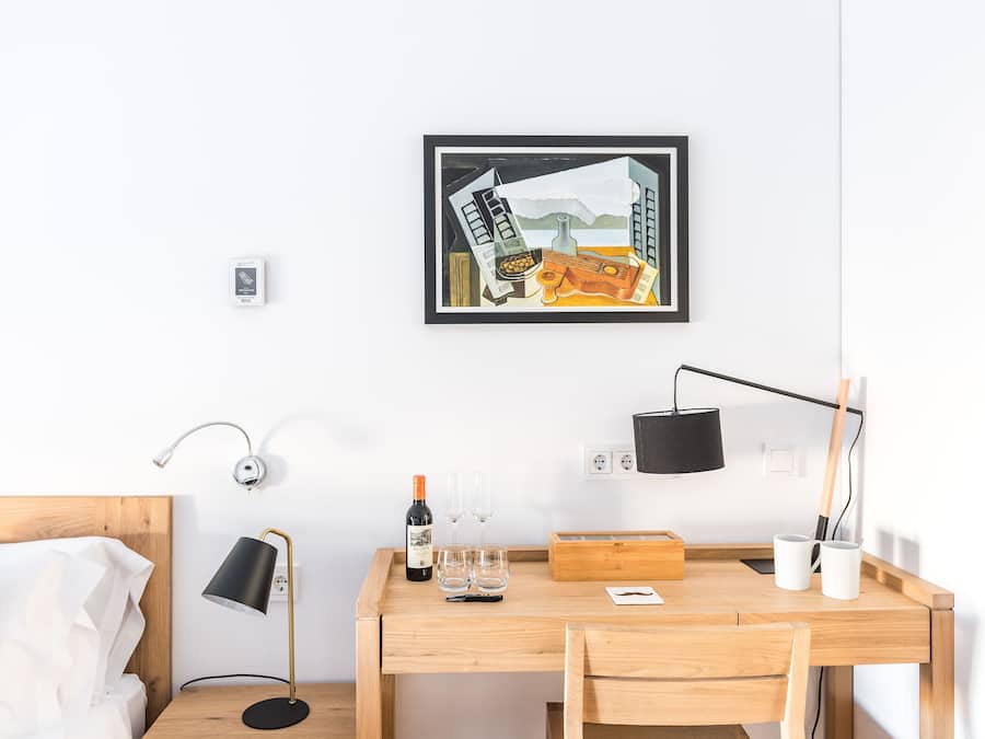 Una habitación de hotel con un escritorio de madera, una cama con ropa blanca, una obra de arte enmarcada en la pared y una mesita de noche con una lámpara.