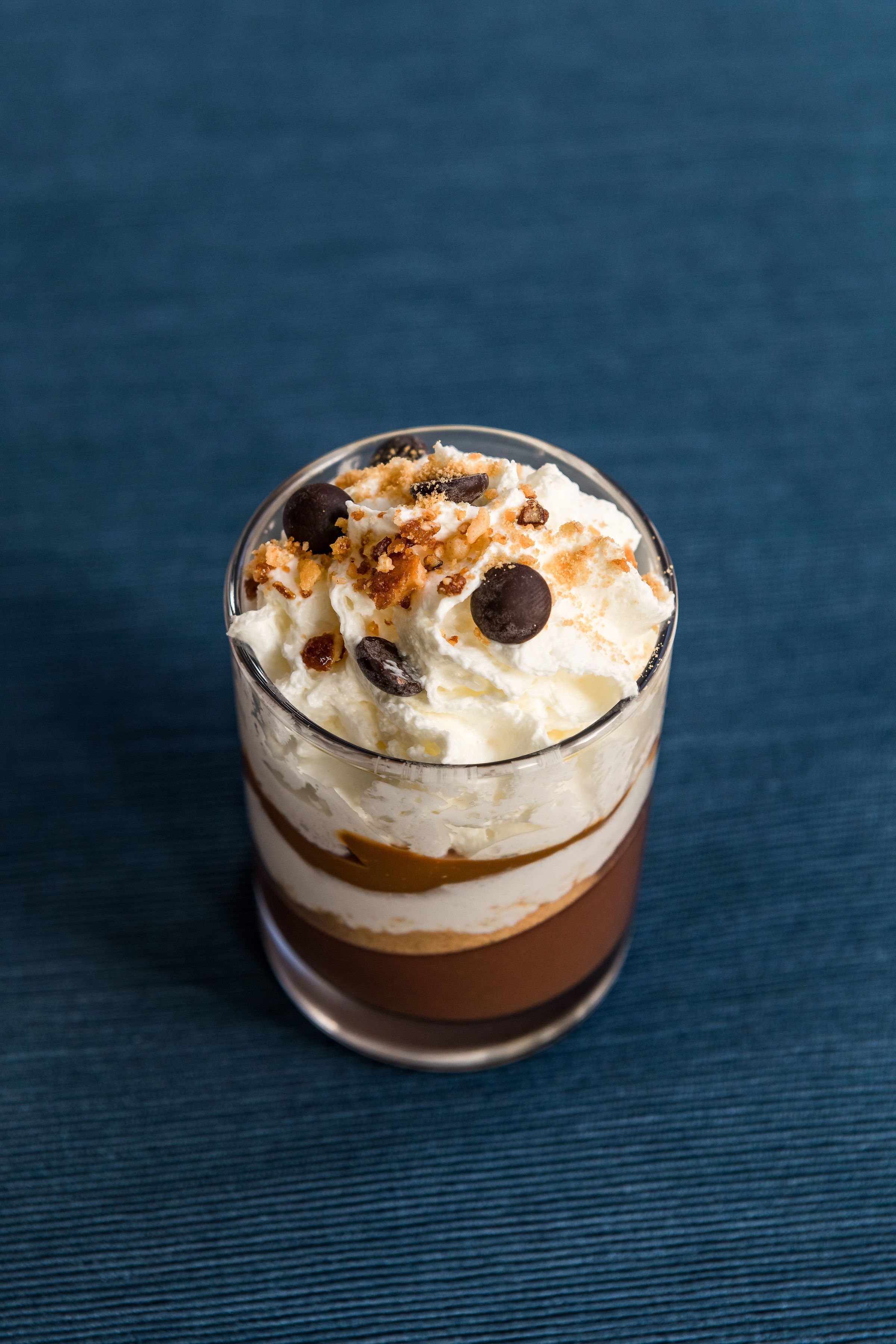A layered dessert in a clear glass on a blue textured surface.