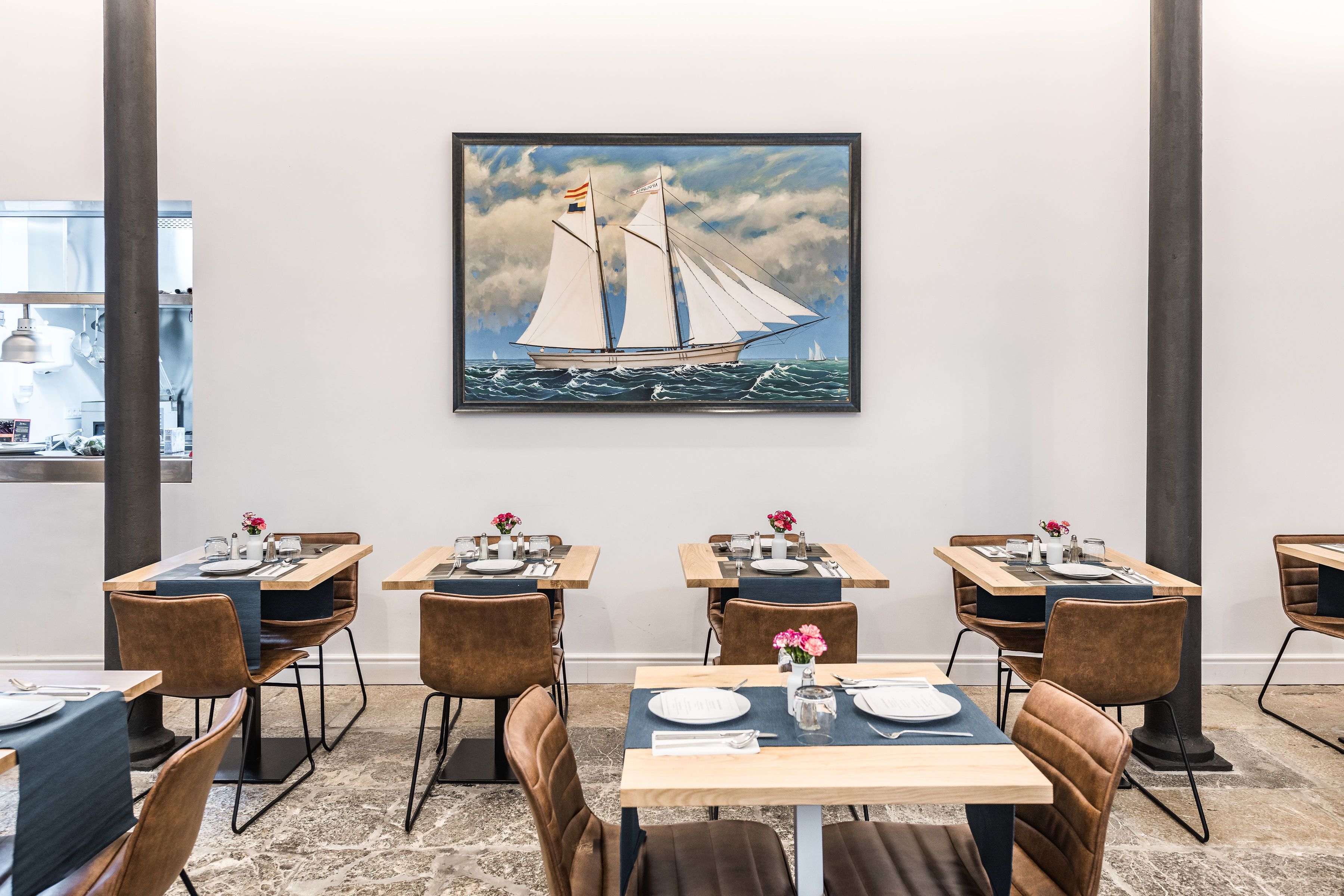 Comedor con mesas dispuestas para cuatro, cada una con un florero, en un interior moderno con un cuadro grande de un velero.