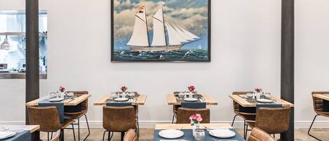 A dining area with tables set for four, each with a vase of flowers, in a modern interior with a large sailboat painting on the wall.