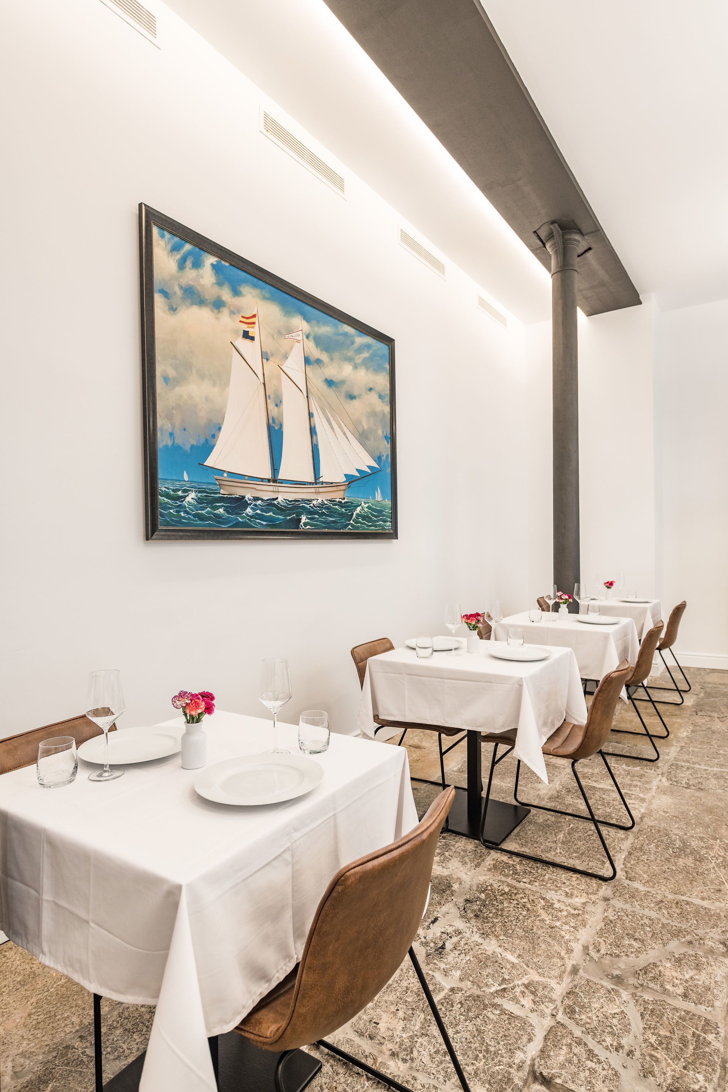 A dining area with tables set for service, a large painting of sailboats on the wall, and a stone floor.