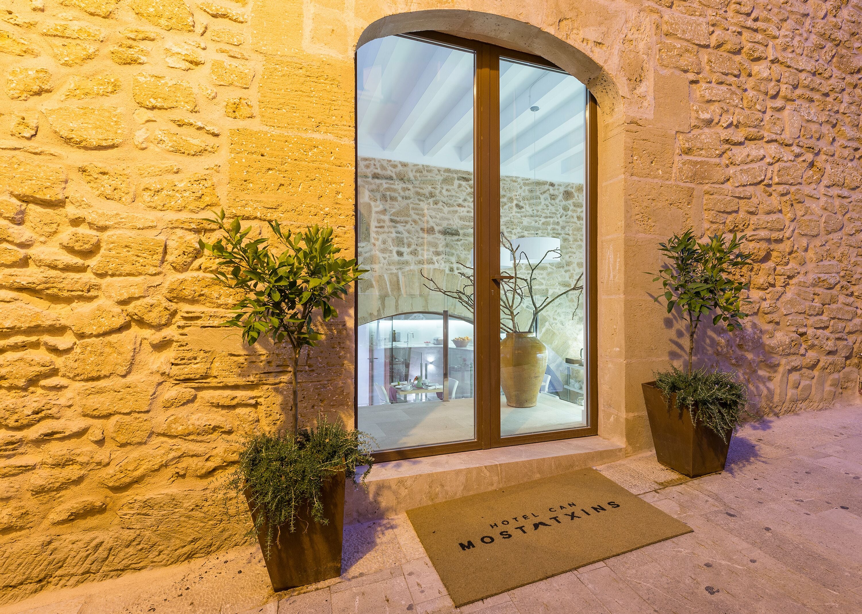 Entrada de un edificio de piedra con puerta de cristal, plantas en macetas y felpudo con el nombre del hotel.