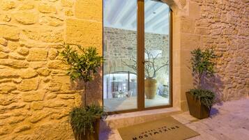 Entrada de un edificio de piedra con puerta de cristal, plantas en macetas y felpudo con el nombre del hotel.