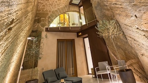 Un interior espacioso con techo alto, paredes de piedra y tragaluces. Hay zonas de estar con cojines y una planta en maceta.