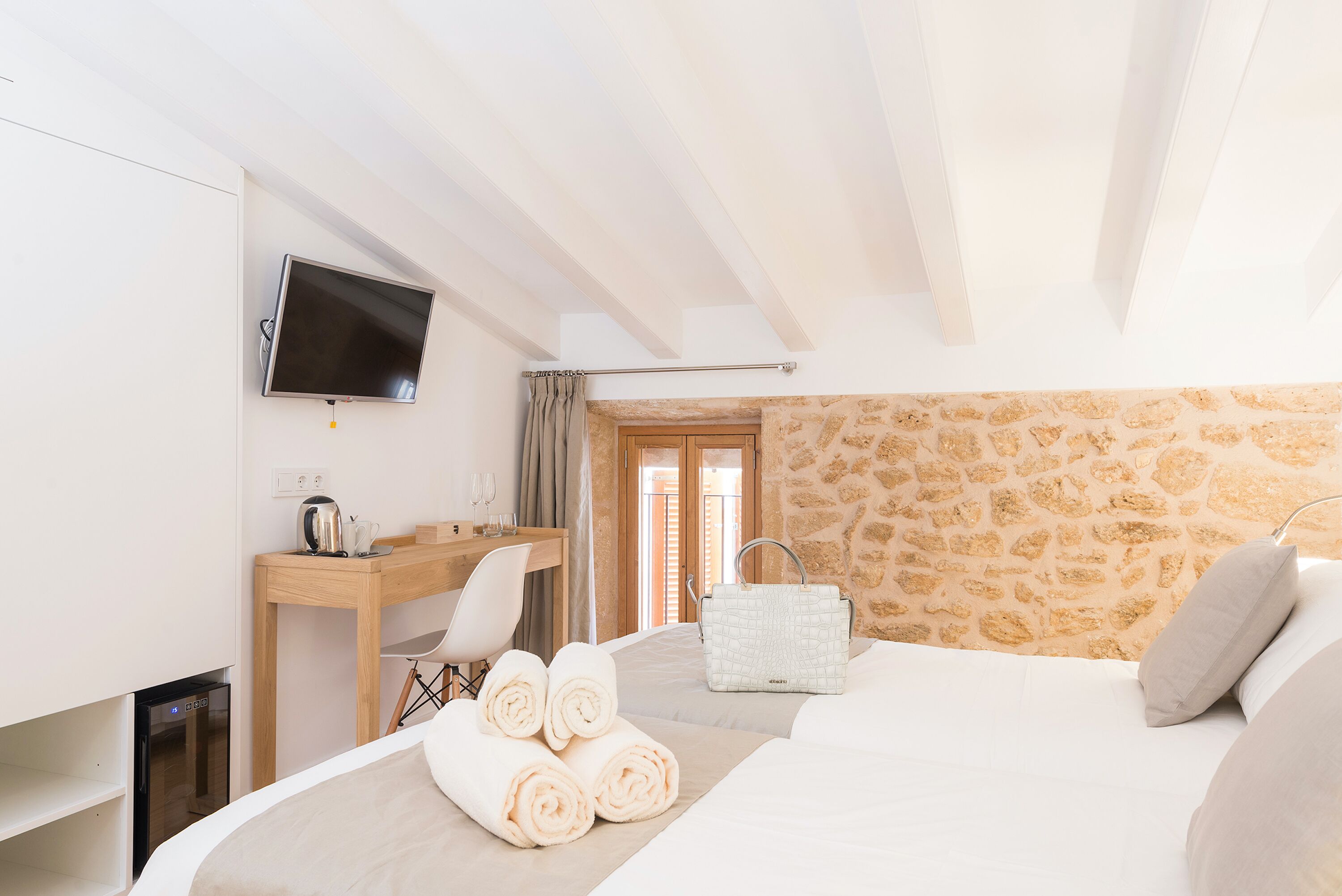 A hotel room with a bed, a desk with a chair, a TV mounted on the wall, and a stone wall.