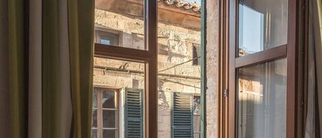 Une pièce avec un plancher en bois, une fenêtre avec des volets verts et une vue sur un vieux bâtiment dont le toit est recouvert de tuiles.