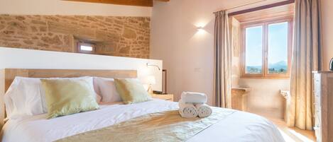 A bedroom with a stone wall, wooden beams, a bed with white linens, a bedside table, and a window with curtains.