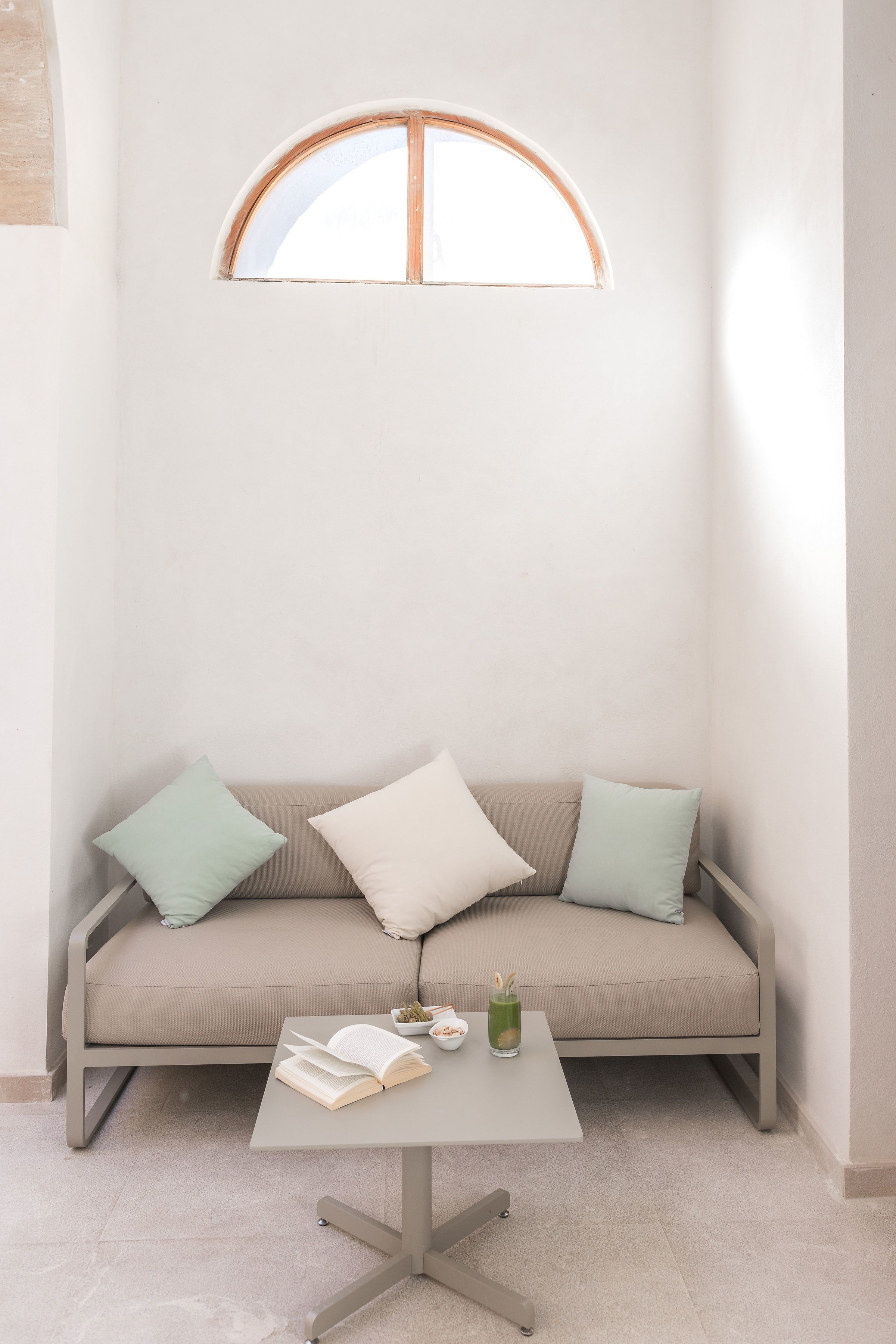Habitación minimalista con sofá color crema, dos almohadas, una mesita con un libro y un vaso, y una ventana con marco de madera.