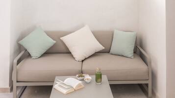 Habitación minimalista con sofá color crema, dos almohadas, una mesita con un libro y un vaso, y una ventana con marco de madera.