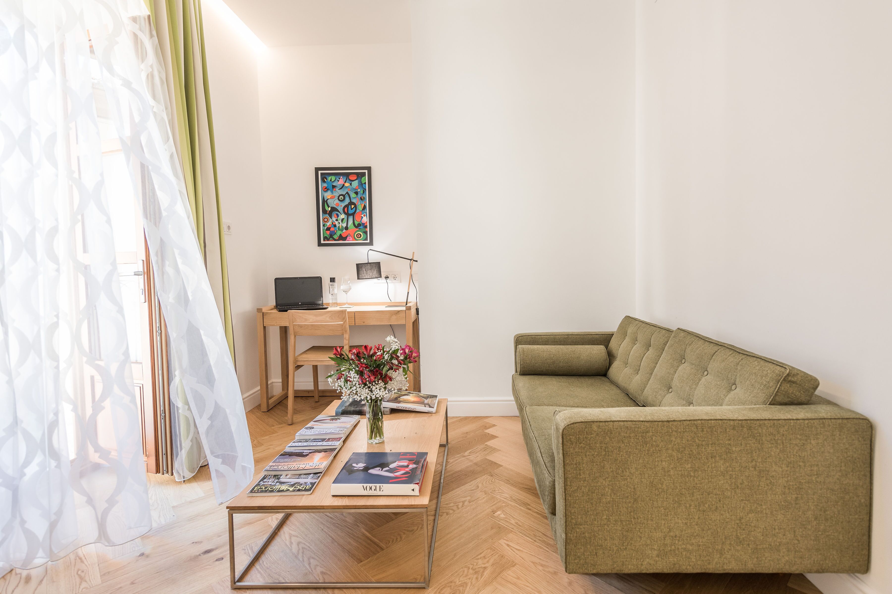 Una sala de estar con un sofá, una mesa de centro con revistas, un escritorio con una computadora y un cuadro en la pared.