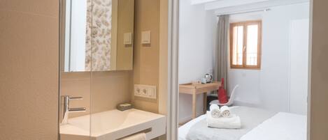 Una habitación de hotel moderna con un espejo grande, un lavamanos blanco, una cama con una colcha blanca y una pequeña mesa con un jarrón rojo.