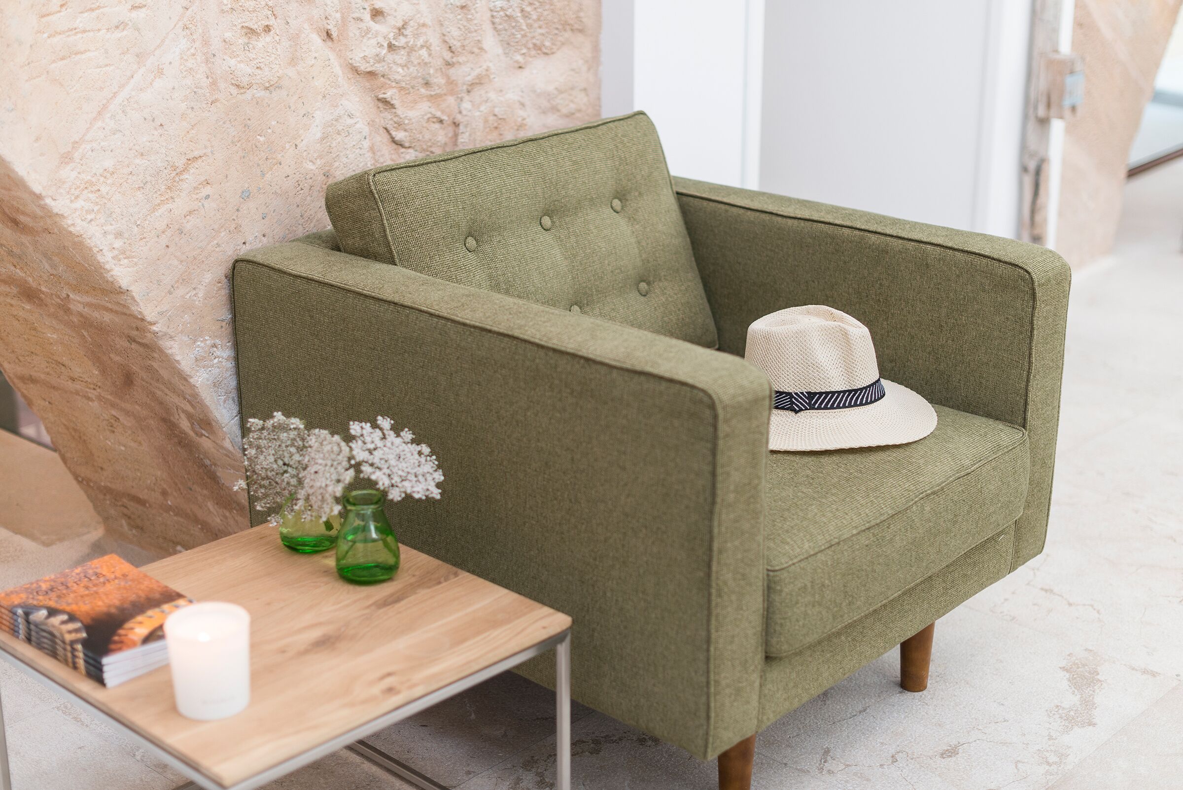 Een groene fauteuil met een witte hoed, een houten tafel met een witte kaars, en een groene vaas met bloemen.