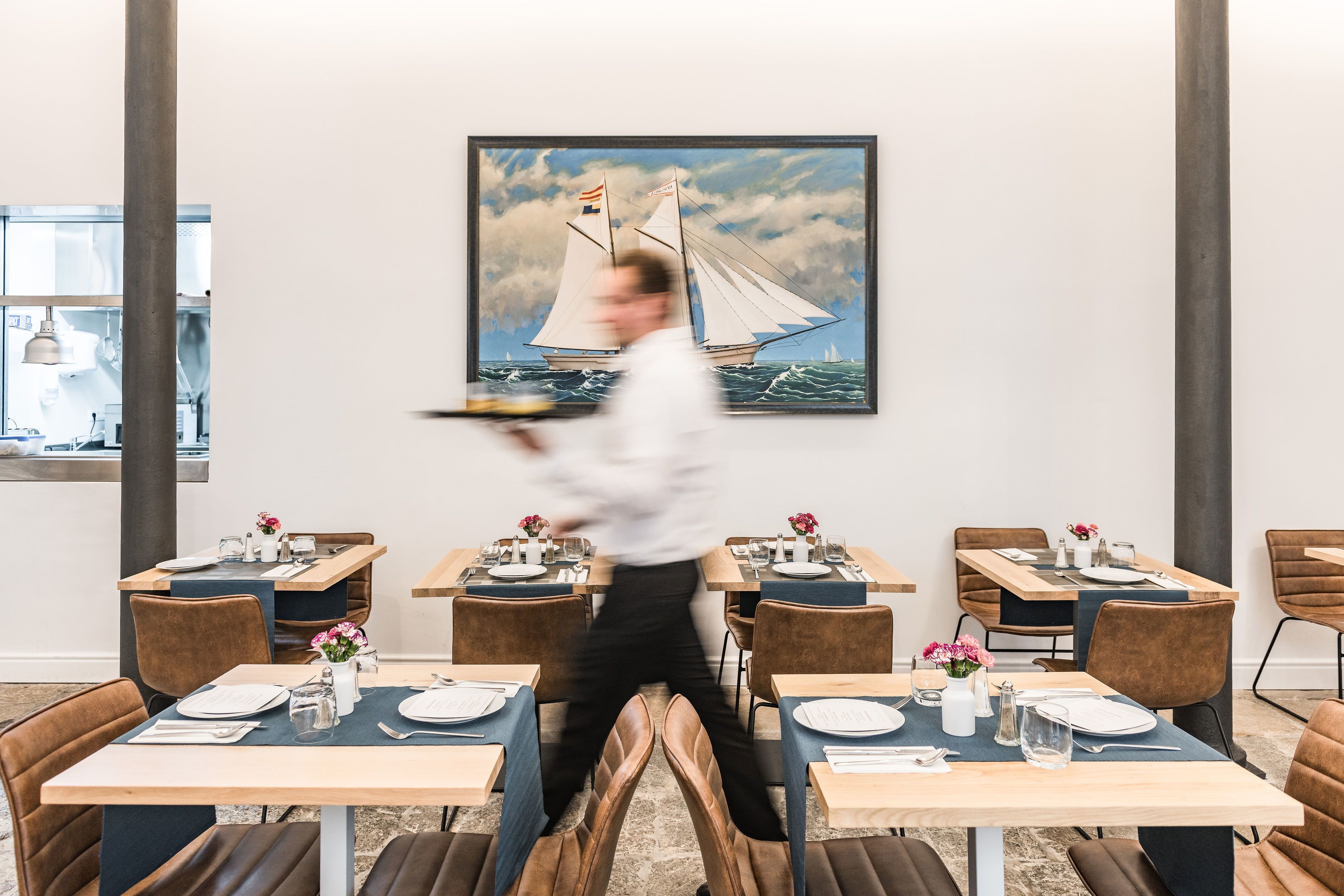 Un restaurant où les tables sont dressées pour le service, un serveur qui s’affaire et un tableau représentant un voilier accroché au mur.
