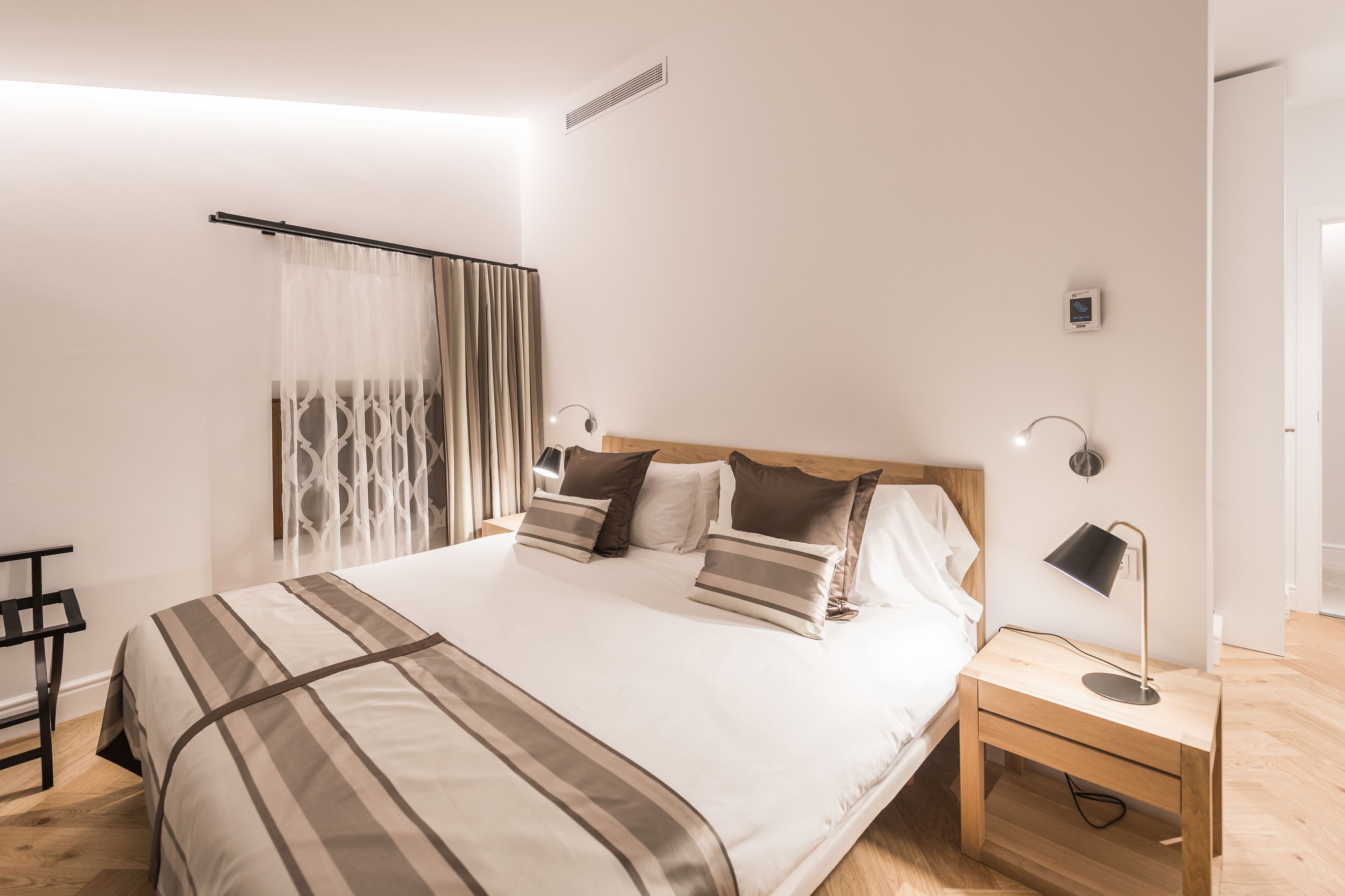 A neatly made bed with striped bedding, a bedside table with a lamp, and a curtain partially drawn.
