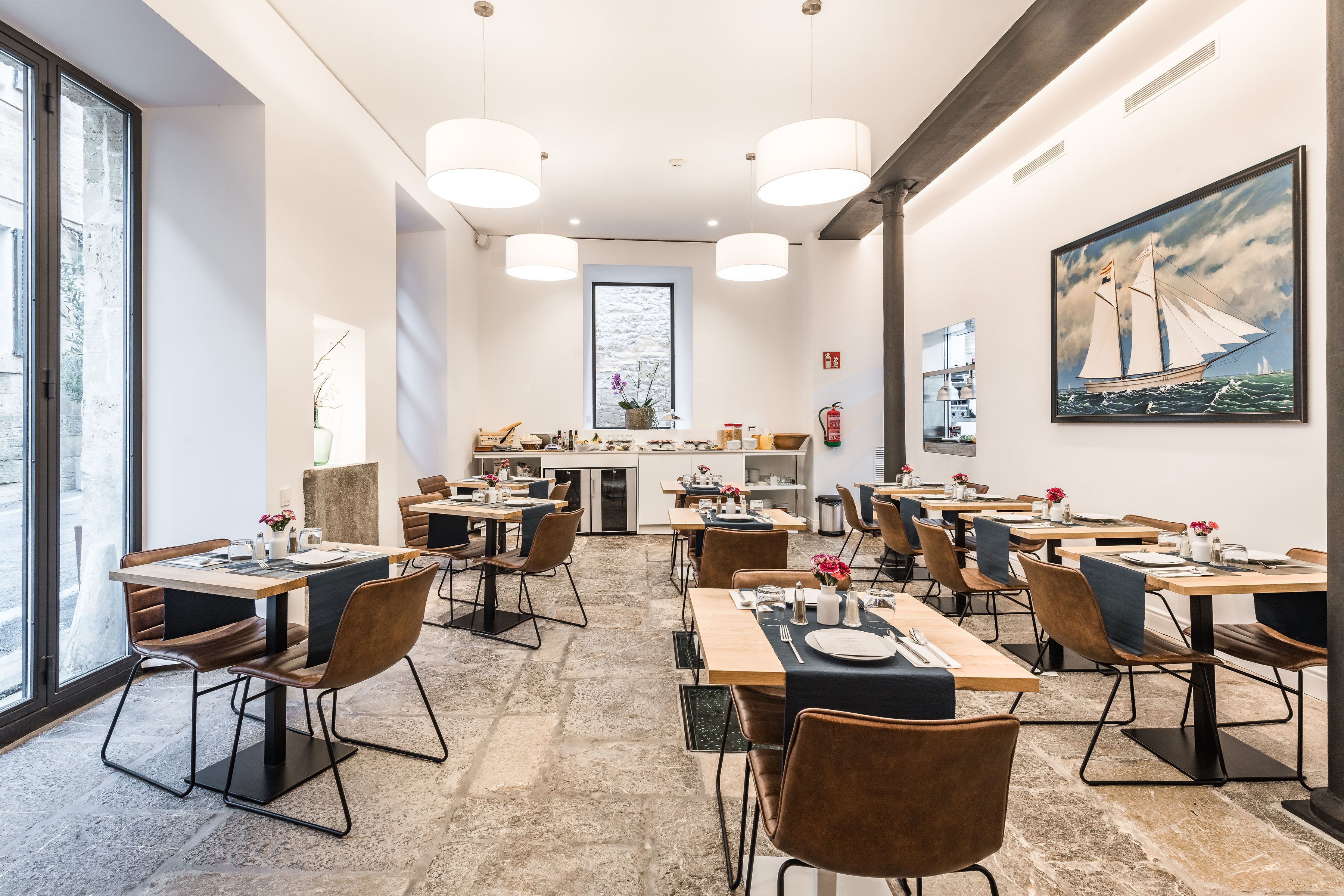 Un espace repas moderne avec des tables et des chaises, une cuisine en arrière-plan et un tableau accroché au mur.