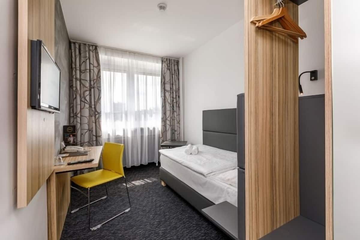 A compact hotel room with a bed, a desk with a chair, a TV, and a coat rack.