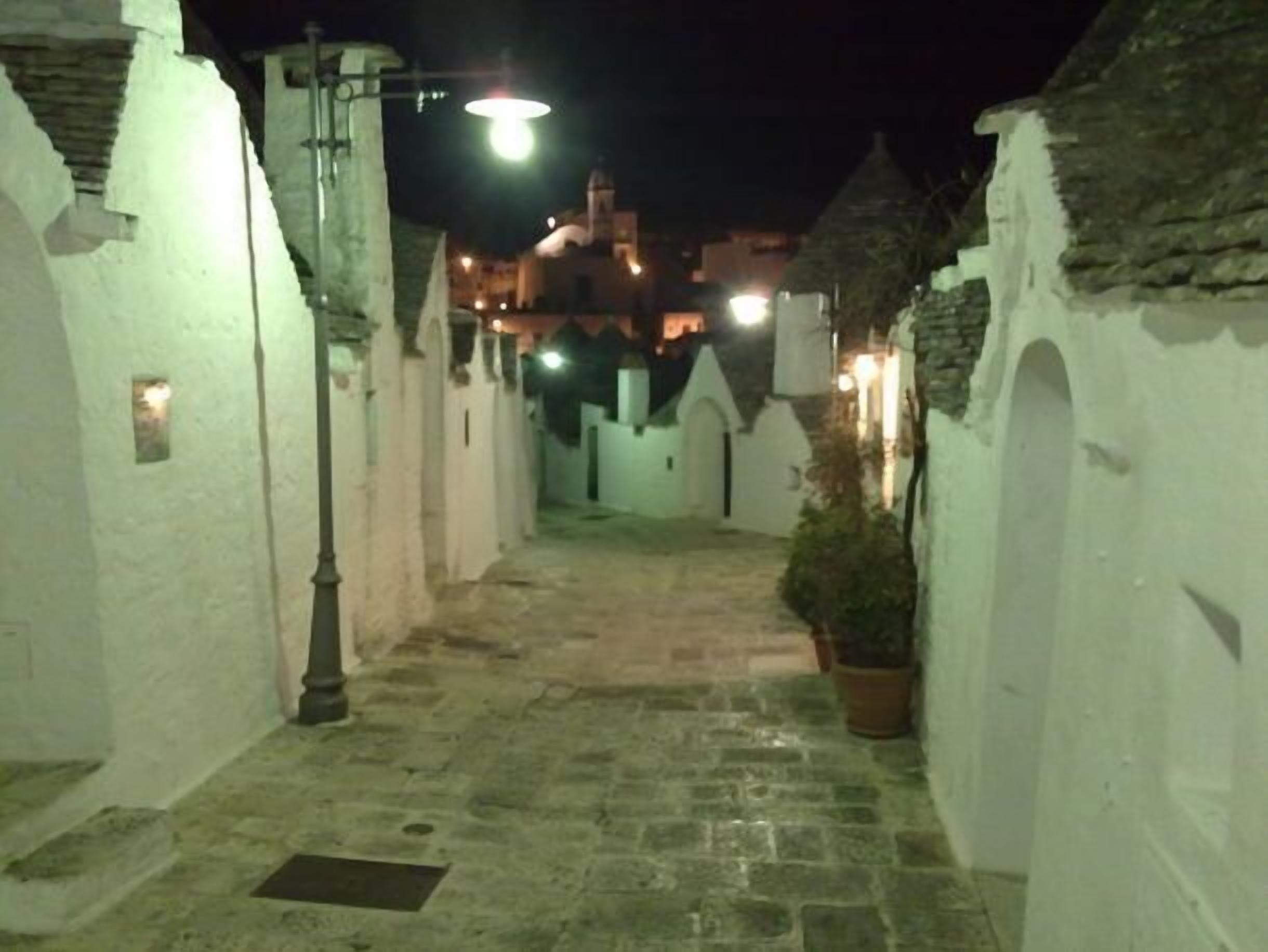 A narrow, stone-paved alleyway with white walls and arched doorways, leading to a building with a dome and illuminated windows.