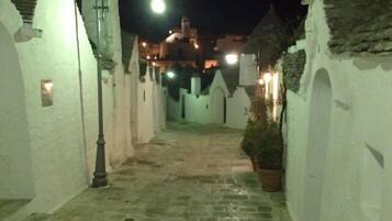 A narrow, stone-paved alleyway with white walls and arched doorways, leading to a building with a dome and illuminated windows.