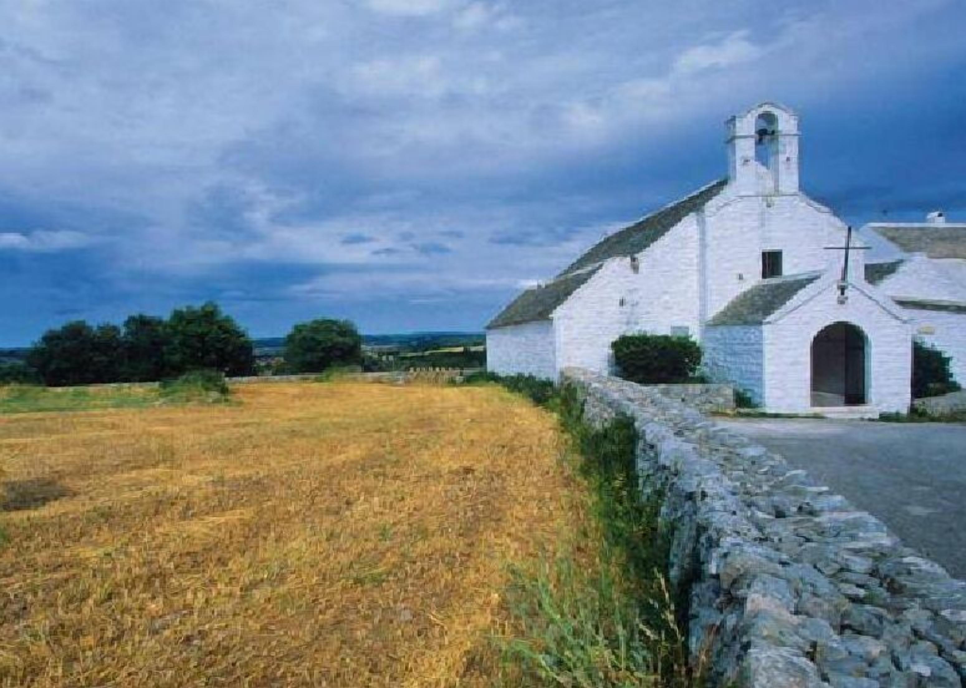 A white church with a bell tower, surrounded by a stone wall and a field of golden crops.