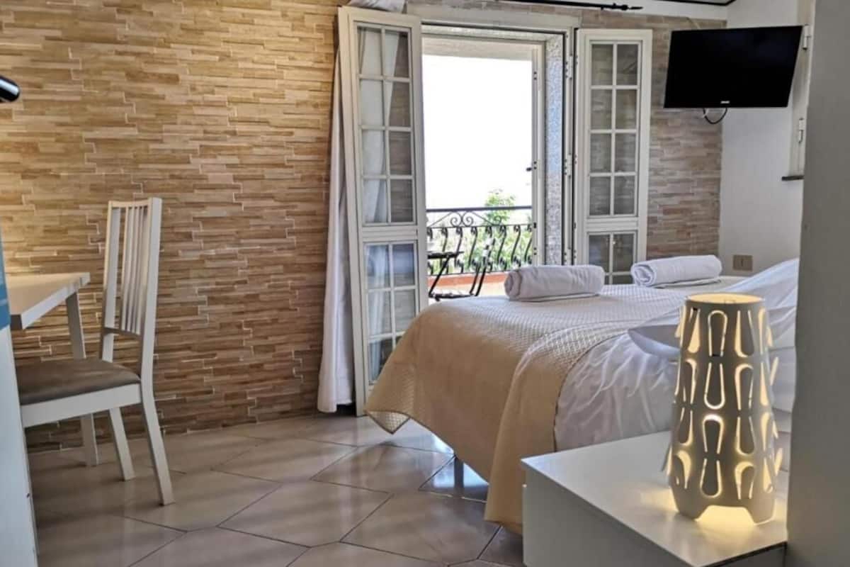 A bedroom with a bed, a chair, a bedside table, a TV, and a balcony view.