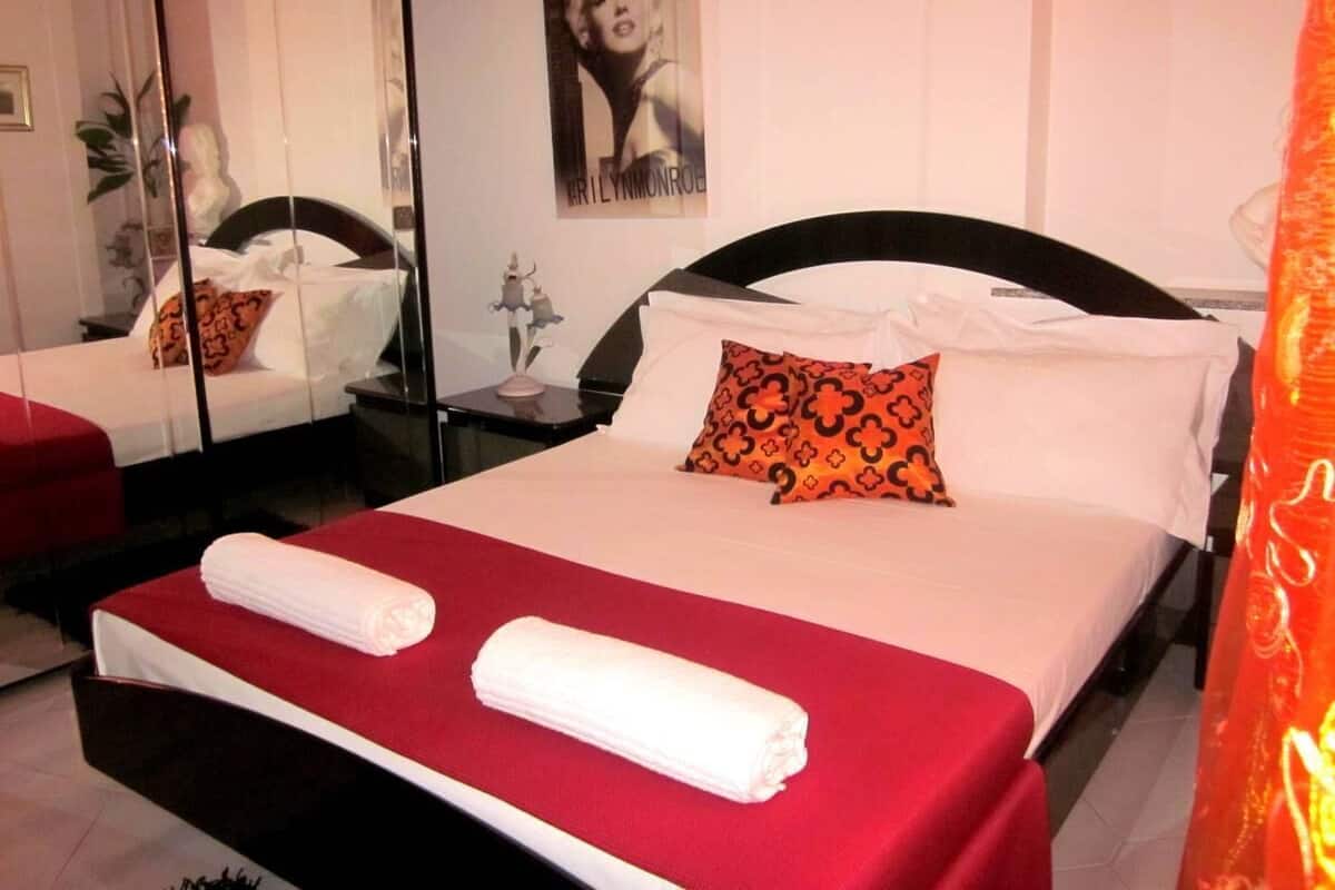 A hotel room with a neatly made bed, a red bedspread, and a large mirror reflecting the room.