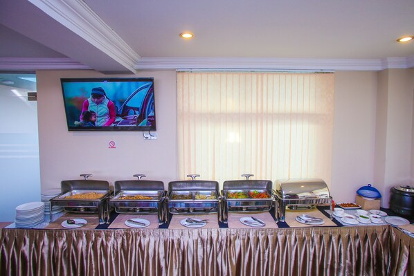 A buffet setup with multiple food stations, a television mounted on the wall, and a window with blinds.